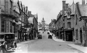 Broad Street, Knighton. This is a postcard loaned by Sally-Anne Richards. Printed on the back is 'Photograph by P.B. Abery, West End Studios, Builth Wells.' The large sign on the left is for 'Williams & Wooding Motor Depot'. Just below and to the left of it is a sign for the 'Norton Arms'. Slightly beyond is a sign for 'Jackson & McCartney's Auction Yard.' In the distance to the left of the Knighton clock tower on the pull-out shop canopy it reads 'H. Gwyther & Sons Boot Stores.' Above it is a sign for the George & Dragon Hotel. Signs on the right of the street include for Bradley's. Above the door which is on the corner on the right is written Wills's Legation and below it there's a sign for 'National Health Insurance Opticians'. On the back of the near van parked on the right is written 'Get It At Allcock's'. Picture is undated but based on the appearance of the old cars may be early 1930s.
