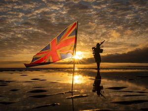 The silhouette of a piper playing on a beach at sunrise, next to a British flag