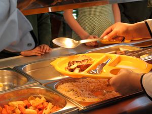 Dinner lady serves up school meals on a yellow plastic tray to primary school pupils waiting at the hot plate in dinner queue