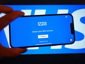 A smartphone displaying the NHS app in front of an NHS sign