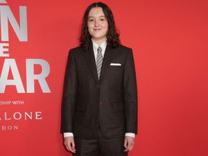 Bella Ramsey, wearing a suit and tie, smiles in front of a red background while attending the GQ Men of the Year photocall at Kensington Roof Gardens in London