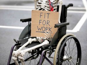 A skeleton sitting in a wheelchair holding up a sign reading ‘Fit for work’