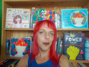 Sarah Savage, wearing a blue vest and with bright red hair, in front of a bookshelf