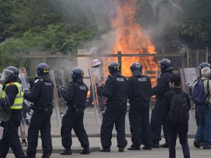 A line of police officers in riot gear, with a fire on wasteland in the background