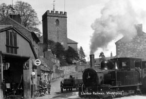 The Llanfair railway at Welshpool, with Welshpool church in the background. An undated image, possibly the 1920s. The circular sign is for 'Horace A. Ballard, Shell Garage.' Railways. Picture-postcard shared by postcard collector Sally Anne Richards.