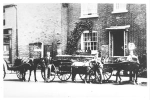 An image of an early milk delivery service, pictured in Albrighton. Dated from the early 20th century, the image shows three milkmen, with Charles Yates the man in the centre. The horses and carts are pictured in front of a substantial house built of brick with sash windows.