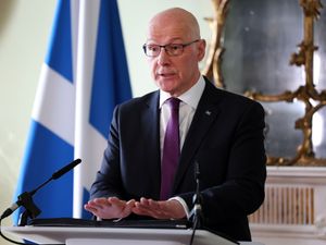 John Swinney speaking from a lectern, with a Scottish flag behind him