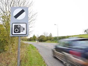 A car passes a national speed limit sign, which also warns of speed cameras on the road ahead