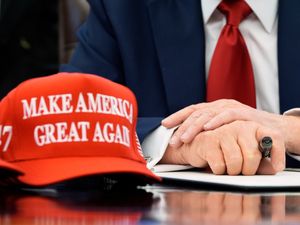 A Make America Great Again red and white baseball cap sits next to Donald Trump's hands holding a pen as they rest on an executive order