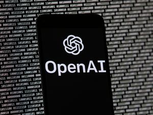 The OpenAI logo on a mobile phone in front of a computer screen displaying random binary data