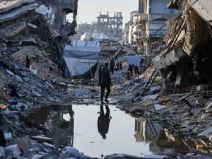 Man, reflected in a puddle, stands in the middle of a street with huge piles of rubble either side of him - everything is destroyed.