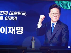 Lee Jae-myung speaking on a stage, overshadowed by a huge image of himself smiling, with one fist raised