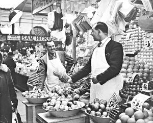 Ronald Gough (right) and his cousin Jim standing at the front of their fruit stall at Old Retail Market Hall, North Street, Wolverhampton, May 1960. Ron is holding out a pineapple to Jim. There is a sign for "Sam Hicks" visible in the background.