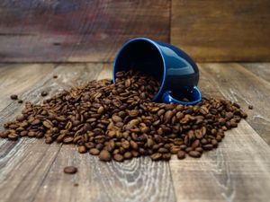 A blue coffee cup is tipped over with beans spilling out of the cup onto a wood surface.