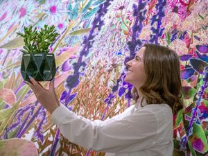 A woman smiling while holding up a plant in front of a large floral mural