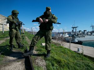 Russian soldiers on an embankment next to a port where naval vessels are moored