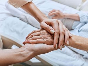 Relative holding trembling hand of senior woman with Parkinson's disease who is lying in hospital bed