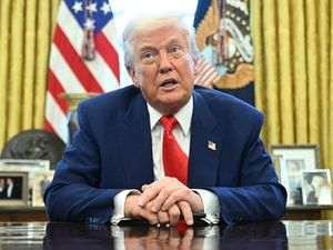 Donald Trump sitting at the Resolute Desk in the Oval Office with his hands clasped in front of him