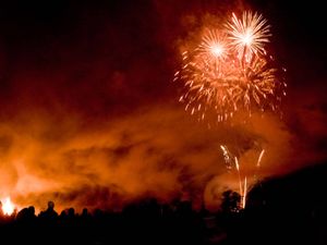 Silhouettes of people standing around a bonfire looking up at fireworks in the night sky