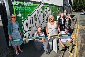 Councillor Linda Leach cuts the ribbon for the mural. With her crouching down is Jenna Wilkins, Nick Chwiej, and with them are supporters and volunteers Genine Riley, Steph Haynes, Marlene Perry, Manny Singh Kang, Michelle Whitehead and Meg Winnington