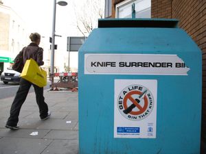 A person walks past a blue Knife Surrender Bin for depositing knives, with a Metropolitan Police sticker on the side reading 'Get A Life, Bin That Knife', in Kings Road, London