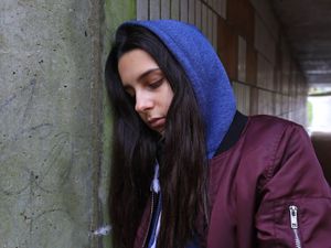 Teenage girl standing alone with a blue hood up and leaning against a wall