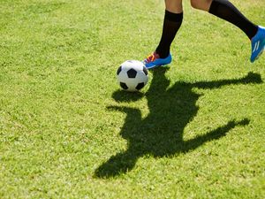 The legs of a person dribbling a football, casting a shadow on a football pitch