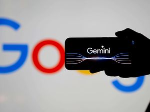 Silhouette of a hand holding a phone with the Gemini logo on its screen with the Google logo partially obscured in the background