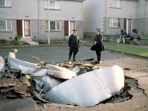 Two police officers standing next to wreckage of a downed plane, partly embedded in a road next to some houses