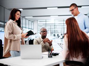 Four colleagues are having a meeting in an office, sitting and standing around a white table with an open plan office in the background