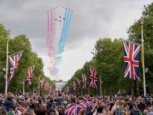Red, white and blue smoke from the back of jets flying in a V formation. Below is a street filled with people and union flags hanging from poles.