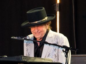 Bob Dylan wearing a black hat and smiling while sitting behind a microphone