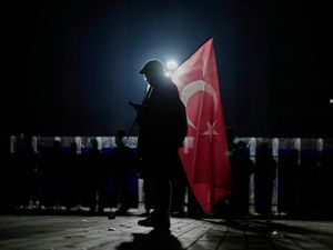 Man holding a Turkish flag over his shoulder is silhouetted by a light behind him. There is a line of riot police with riot shields behind him