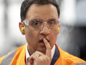 Headshot of Anas Sarwar wearing protective glasses and ear buds, with a finger resting on his mouth