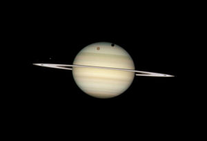 A picture of the four moons of Saturn passing in front of their parent planet taken by Hubble. In this view, the giant orange moon Titan casts a large shadow onto Saturn's north polar hood. Below Titan, near the ring plane and to the left is the moon Mimas, casting a much smaller shadow onto Saturn's equatorial cloud tops.