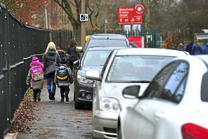 Youngsters and a parent squeeze past cars parked on the pavement near the school